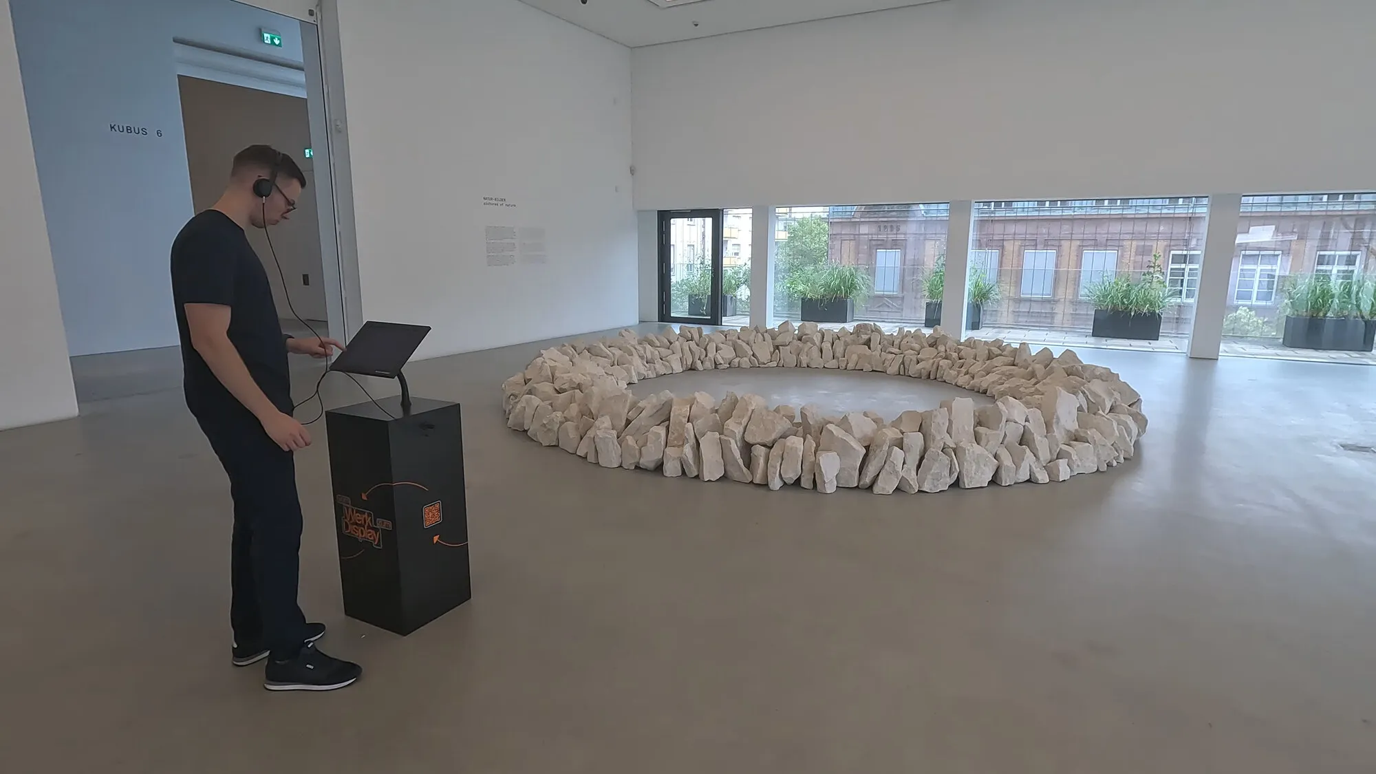 An exhibition kiosk with headphones in a hall of Kunsthalle Mannheim, beside Richard Long’s stone circle.